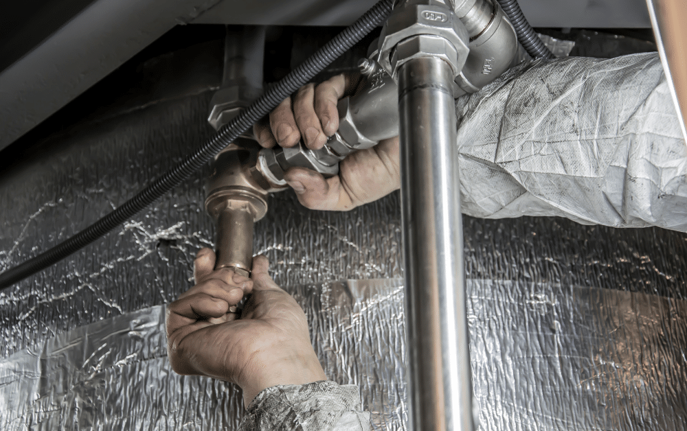 Hands connecting metal pipe fittings and valves under an insulated ceiling, with cables and a vertical stainless steel pipe nearby