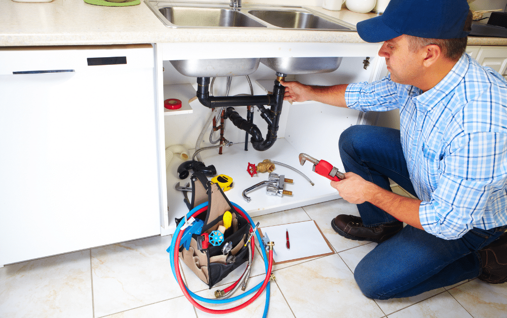 Plumber kneeling under double kitchen sink, tightening drain pipes with wrench; tools and hoses on floor in open cabinet.